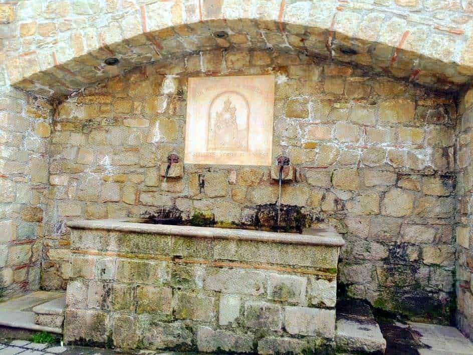 Fonte di acqua in pietra con le due rubinetterie, sullo sfondo un affresco sacro, ambiente storico in pietra con volta a botte, perfetto esempio di fontana antica in stile rustico.
