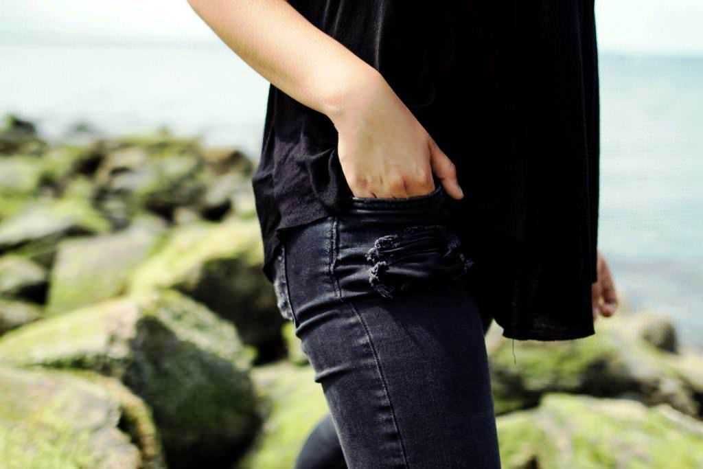Vestito casual donna con jeans strappati e giacca nera, stile streetwear, ambientazione all'aperto sulla spiaggia.