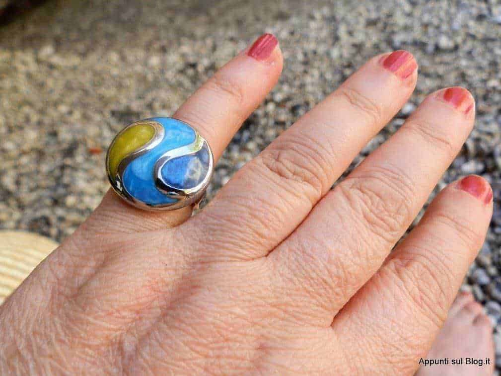 Anello in metallo con pietra colorata blu e verde, indossato su una mano femminile con unghie smaltate di rosso, sfondo di ciottoli e superficie naturale.