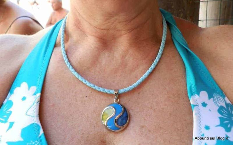 Orecchini e collana artigianali con farfalle colorate, esposte su un contenitore di vimini giallo e posizionate su un tovagliolo con fiori, accanto a una donna sorridente che indossa gli stessi gioielli, con occhiali scuri e sciarpa in testa.