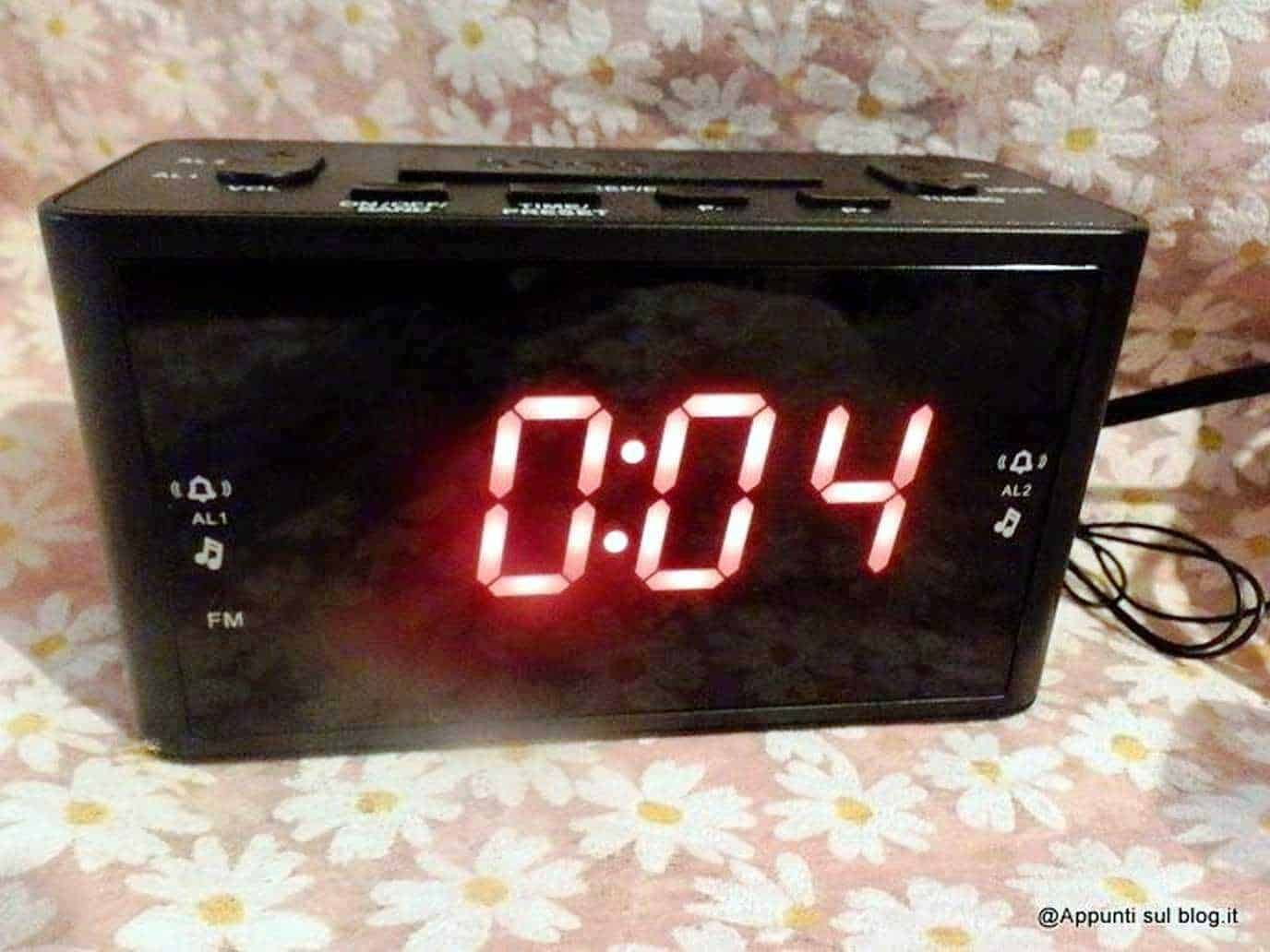 Orologio digitale nero con display rosso che mostra le 7:04, su sfondo floreale rosa e bianco.