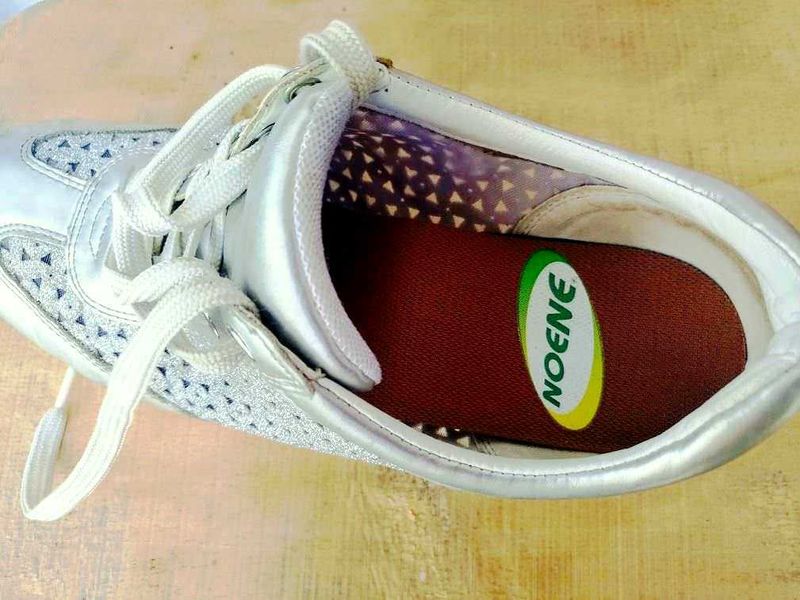 Solette antishock Noene in carbonio per ogni esigenza sportiva. 7 Scarpa da ginnastica bianca con dettagli colorati, vista dall'alto su superficie di legno. ideata per sport e moda casual, offre comfort e stile quotidiano.