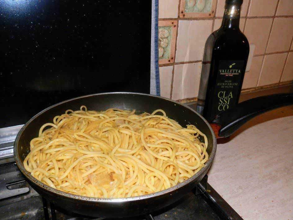 Cottura di spaghetti italiani in padella con olio extravergine di oliva sulla cucina domestica. Bottiglia di olio e piano di lavoro tisano in ceramica sullo sfondo.