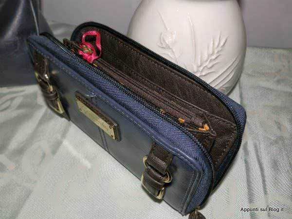 Borsa messenger blu scuro con tasche e zip, accessori e dettagli in pelle, poggiata su una superficie in tessuto, sfondo con un vaso bianco, immagine di oggetti di moda e stile casual uomo/donna.