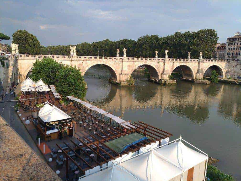 Panorama del Ponte di Castel Sant'Angelo a Roma con locale esterno sul fiume Tevere, ambientazione estiva, tavoli all'aperto, atmosfera urbana e storica, vista sull'acqua e sul ponte con statue decorative, luogo ideale per ristorazione e relax.