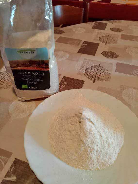 Farina biologica per pizza su piatto bianco, con sacchetto di farina integrale organica sul tavolo decorato con motivi foglia e quadrati, ambiente casalingo, fotografia di alimenti e prodotti biologici.
