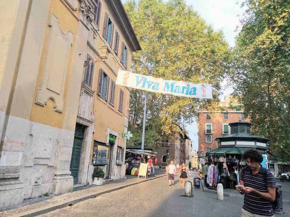 Viva Maria banner appeso tra gli edifici in una strada di Roma, con negozi e venditori ambulanti, atmosfera vivace e al centro una persona che usa il cellulare, aree urbane e vita cittadina italiana.