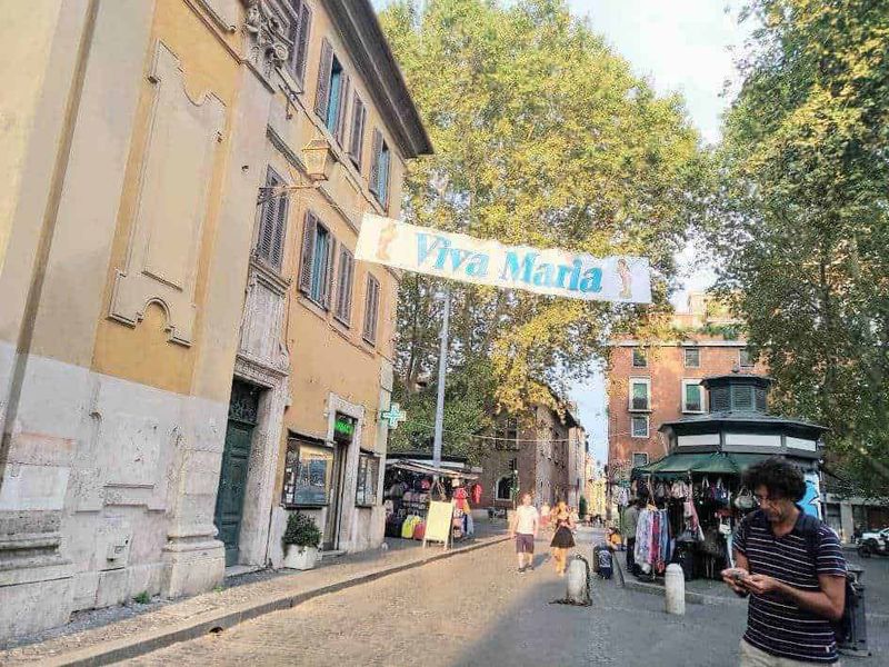 Roma capitale tour in mesi caldi 6 Viva Maria banner appeso tra gli edifici in una strada di Roma, con negozi e venditori ambulanti, atmosfera vivace e al centro una persona che usa il cellulare, aree urbane e vita cittadina italiana.