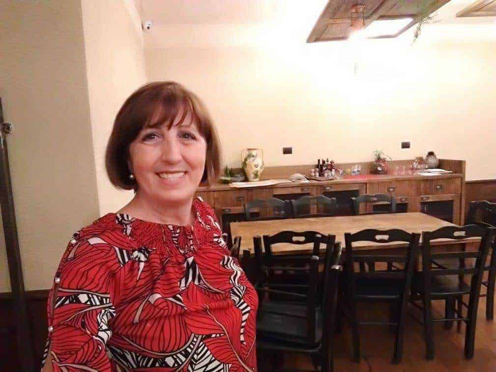 Una donna sorridente in un ristorante accogliente con tavoli e sedie di legno, decorazioni eleganti e atmosfera calda, perfetta per eventi e incontri sociali.