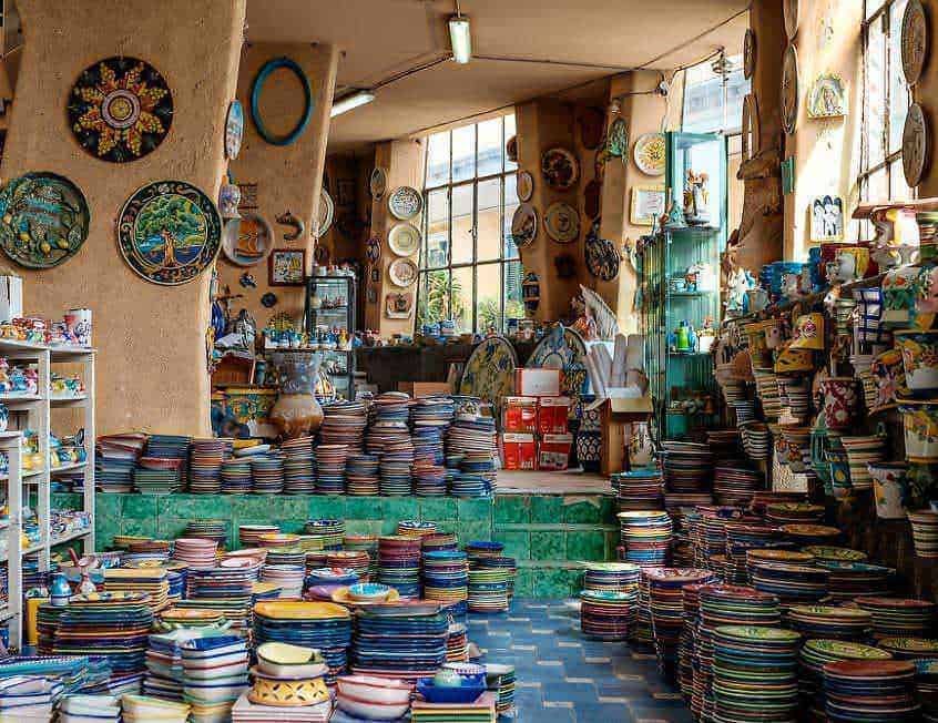 Colorato negozio di ceramiche artigianali, con piatti e oggetti decorativi disposti su scaffali e pavimento, luce naturale che entra dalle grandi finestre, atmosfera accogliente e creativa.
