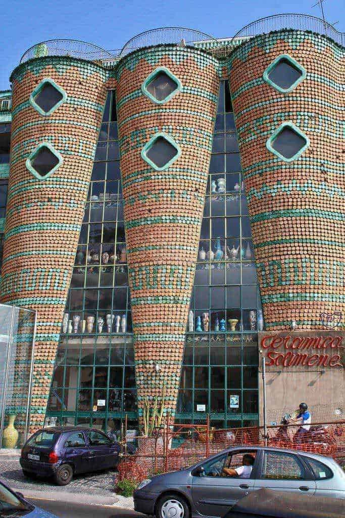 Una facciata di un edificio moderno decorata con mosaici di ceramiche colorate, con grandi vasi di terracotta e vetrate che riflettono il cielo. La struttura si chiama "Ceramica Ramena" ed è un esempio di architettura innovativa e arte urbana.