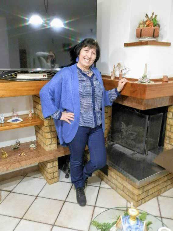 Una donna sorridente con abbigliamento blu posiziona davanti a un camino in un soggiorno accogliente. La scena include decorazioni natalizie e un ambiente casalingo e caloroso.