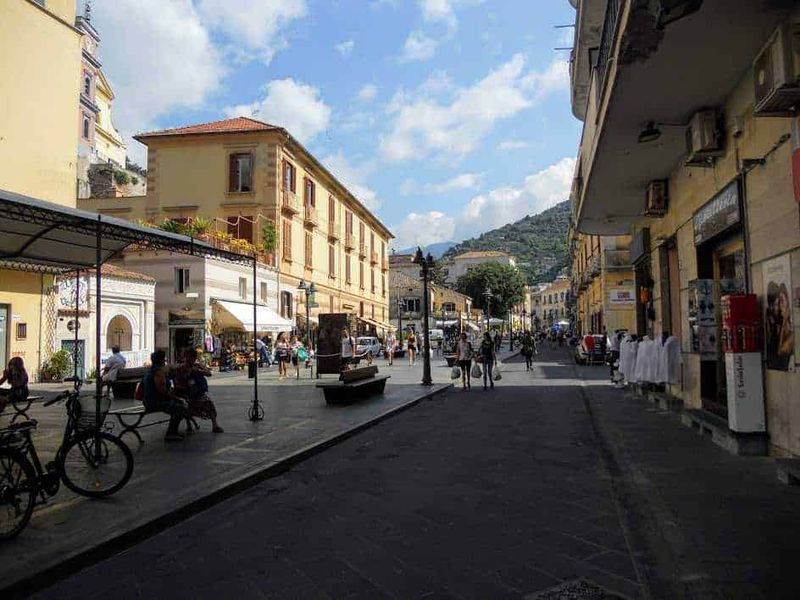 Costiera amalfitana, 18 borghi da visitare in 40 km 12 Vicolo pedonale nel centro storico di Maiori, un caratteristico paese italiano con edificio antichi, negozi, bar e persone che passeggiano sotto un cielo sereno e nuvoloso, con colline sullo sfondo.