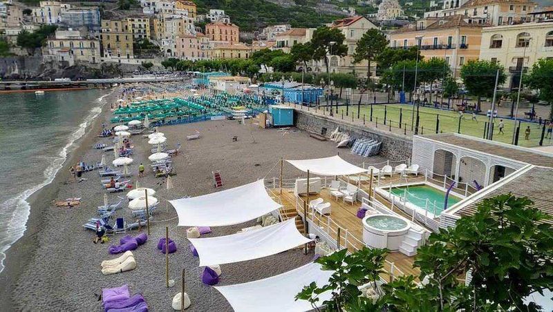 MINORI con spiaggia con lettini e ombrelloni, mare calmo e città sullo sfondo, area relax con piscina, ambientazione estiva e turistica, zona balneare, Italia, 