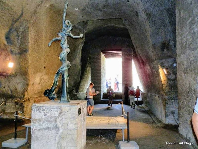 Vita di un uomo con la statua di bronzo in una grotta storica, visitatori che esplorano l'ambiente rupestre e l'ingresso luminoso nel sito archeologico, ambiente suggestivo e ricco di storia.