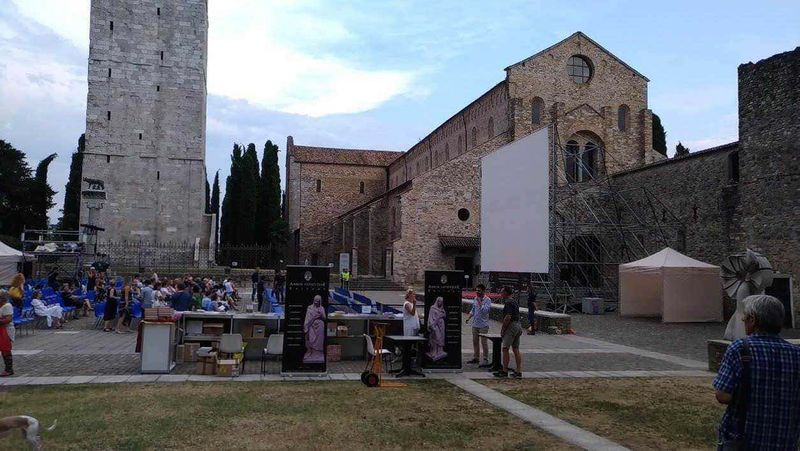 Friuli Venezia Giulia tour nei borghi e province della terra di confine 9 Cinema all'aperto in una storica piazza con antiche mura e torre, partecipanti che aspettano l'inizio della proiezione sotto un cielo nuvoloso, spazio di eventi culturali e sociali.