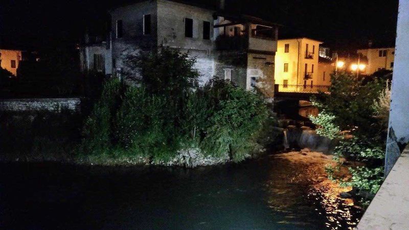 Friuli Venezia Giulia tour nei borghi e province della terra di confine 12 Tratto di fiume urbano di notte, illuminato da luci calde, con edifici antichi sullo sfondo e vegetazione lungo le rive, rappresenta un paesaggio suggestivo tipico di un ambiente cittadino.