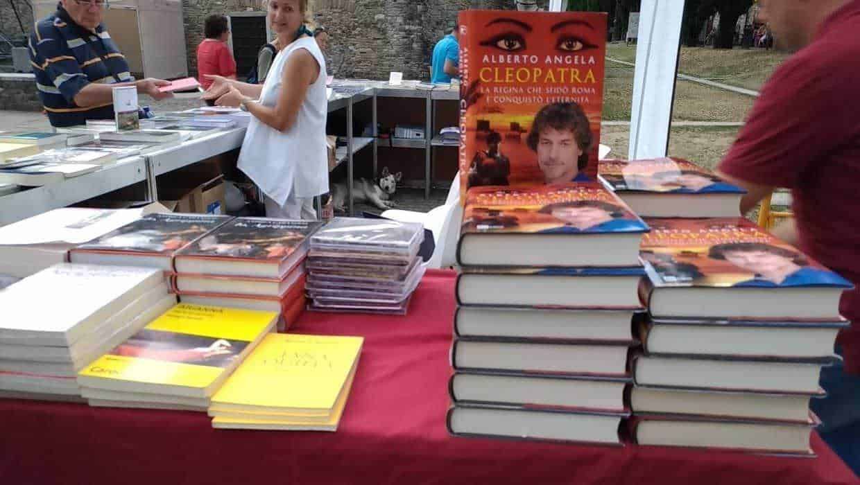 Alto testo only (max 200 caratteri): Bancarella di libri durante una fiera letteraria con copie di Alberto Angela Cleopatra in primo piano, pubblico e stand sullo sfondo.
