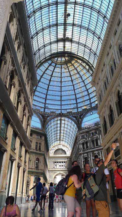 Vetrate decorative in galleria commerciale con architettura storica e turisti che esplorano l'interno di un grande centro commerciale.