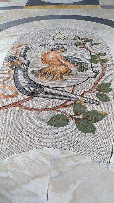 Piastrelle mosaico con scudo e simboli storici, decorazione artistica su pavimento in stile classico, dettagli intricati in mosaico, elemento decorativo nel contesto urbano, esempio di arte decorativa italiana.