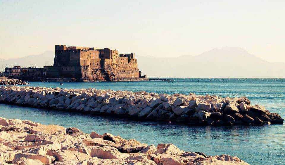 Fortificazione storica sulla costa mediterranea con mare calmo e rocce, vista panoramica al tramonto con montagne sullo sfondo, riflessi dell'acqua e paesaggio costiero suggestivo.