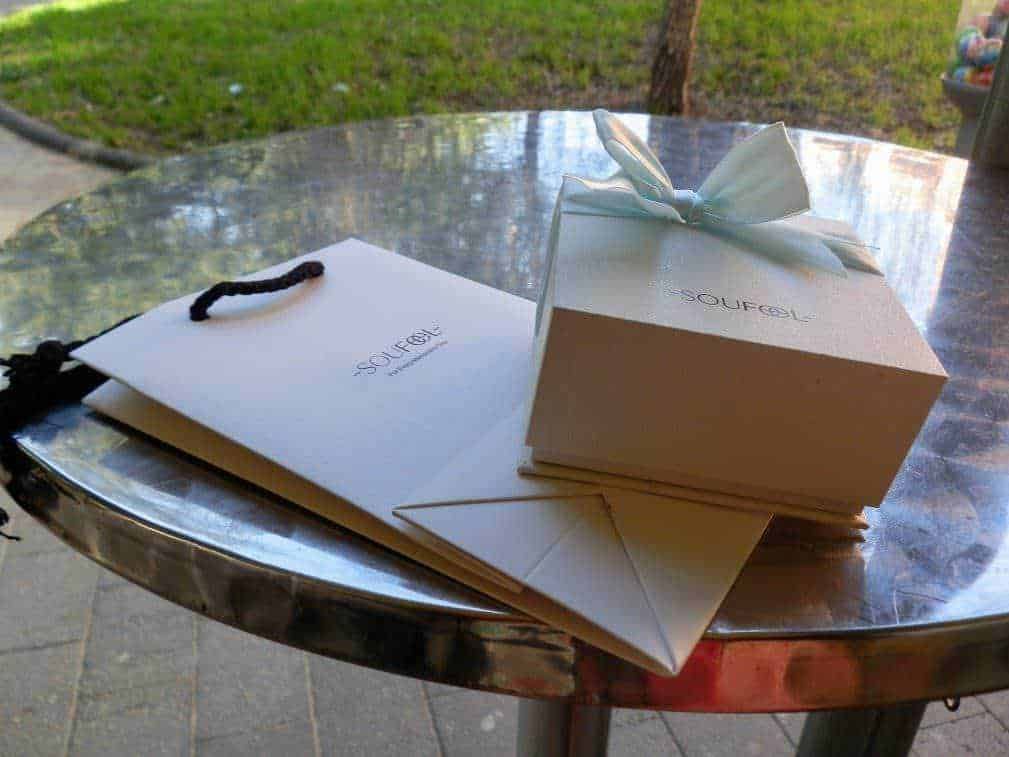 Elegante confezione regalo con fiocco azzurro e elegante shopping bag bianca con logo "SOUFOL" su tavolo in esterno, perfetta per occasioni speciali e regali esclusivi.