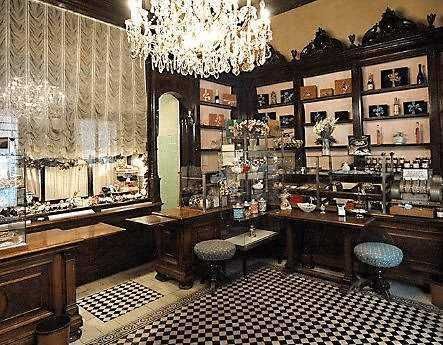 Una stanza elegante con scaffali di legno scuro pieni di oggetti d'antiquariato e decorazioni vintage, illuminata da un lampadario di cristallo, con tende di pizzo e un pavimento a scacchi.