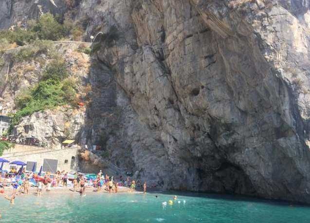 Fronte di spiaggia con scoglio e persone che nuotano e prendono il sole, vista dal mare, in una località balneare italiana.