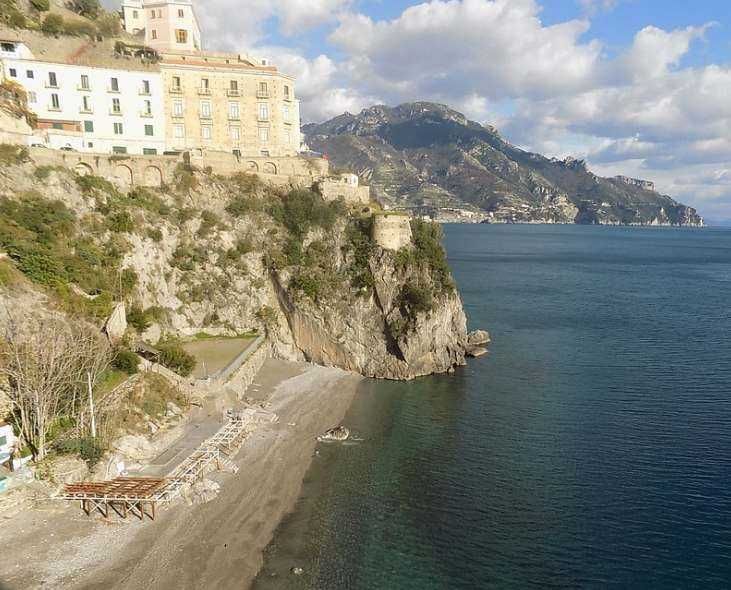 Lungomare di Amalfi con hotel e scogliera, vista mare e montagne, estate, costa italiana, turismo, paesaggio marino, scogliera rocciosa, natura e relax, immagini di viaggio, promozione destinazioni turistiche.