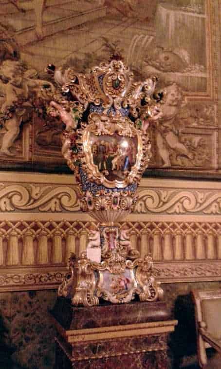 Ornamento religioso barocco dorato con decorazioni intricate e cristalli, posizionato su un pianale in marmo, in un ambiente storico riccamente decorato.