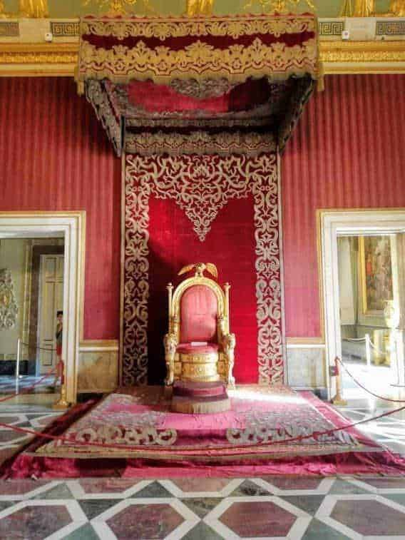 Regina nel trono dorato in uno stile barocco, decorato con ricami e velluto rosso, all’interno di un palazzo storico con pareti elaborate e pavimento a scacchi.