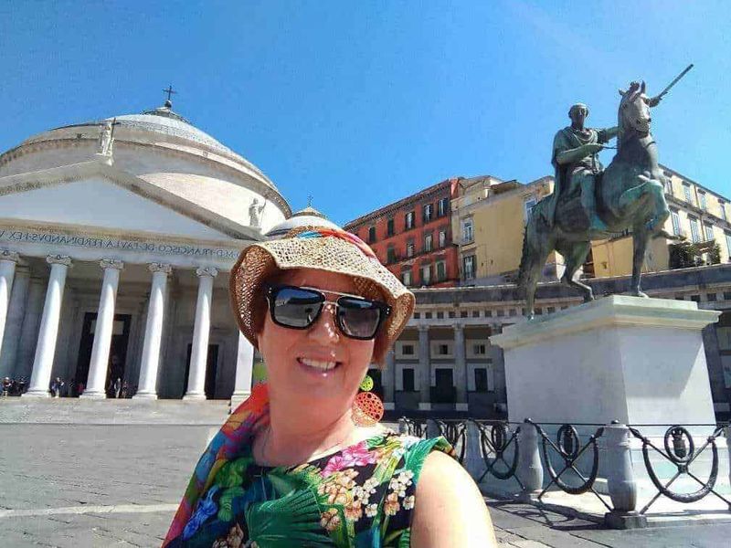 Coppia di monumenti storici a Napoli con una donna sorridente in primo piano, abbigliamento colorato e cappello di paglia, sullo sfondo chiese e statue classiche sotto un cielo azzurro.