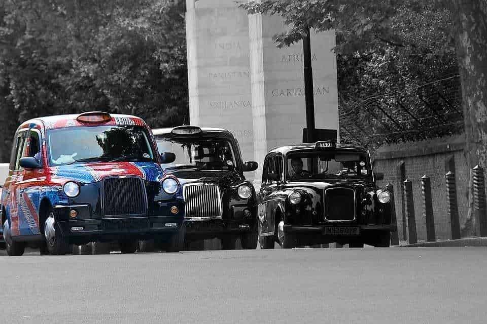 Auto di servizio con livrea colorata e due taxi neri storici parcheggiati lungo una strada cittadina. Scatto in bianco e nero con la vettura di servizio colorata in evidenza.