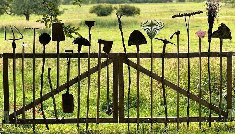 Palette di attrezzi da giardinaggio appesi a una staccionata di legno in un campo verde, ideale per articoli su strumenti, giardinaggio e coltivazione sostenibile.