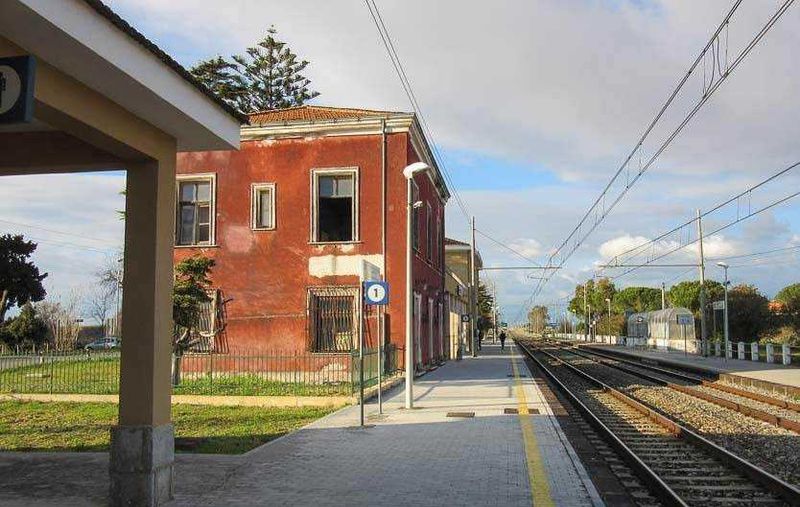 Edificio storico rosso lungo la stazione ferroviaria sul binario, con piattaforma pedonale, linee elettriche aeree e paesaggio naturale circostante, ideal per foto di viaggio e cultura locale.