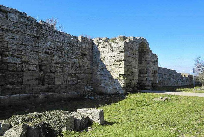 Muri romani antichi in pietra, resti di un sito archeologico storico in Italia, con un cielo blu e un'area verde circostante. Aperto al pubblico e ideale per appassionati di storia e archeologia.