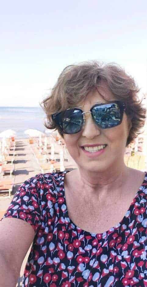 Fotocamera di una donna con occhiali da sole sulla spiaggia, mare e pedane con sedie e ombrelloni in sfondo, estate e vacanze al mare, ritratto di donna sorridente in abbigliamento estivo, viaggio e relax al sole.