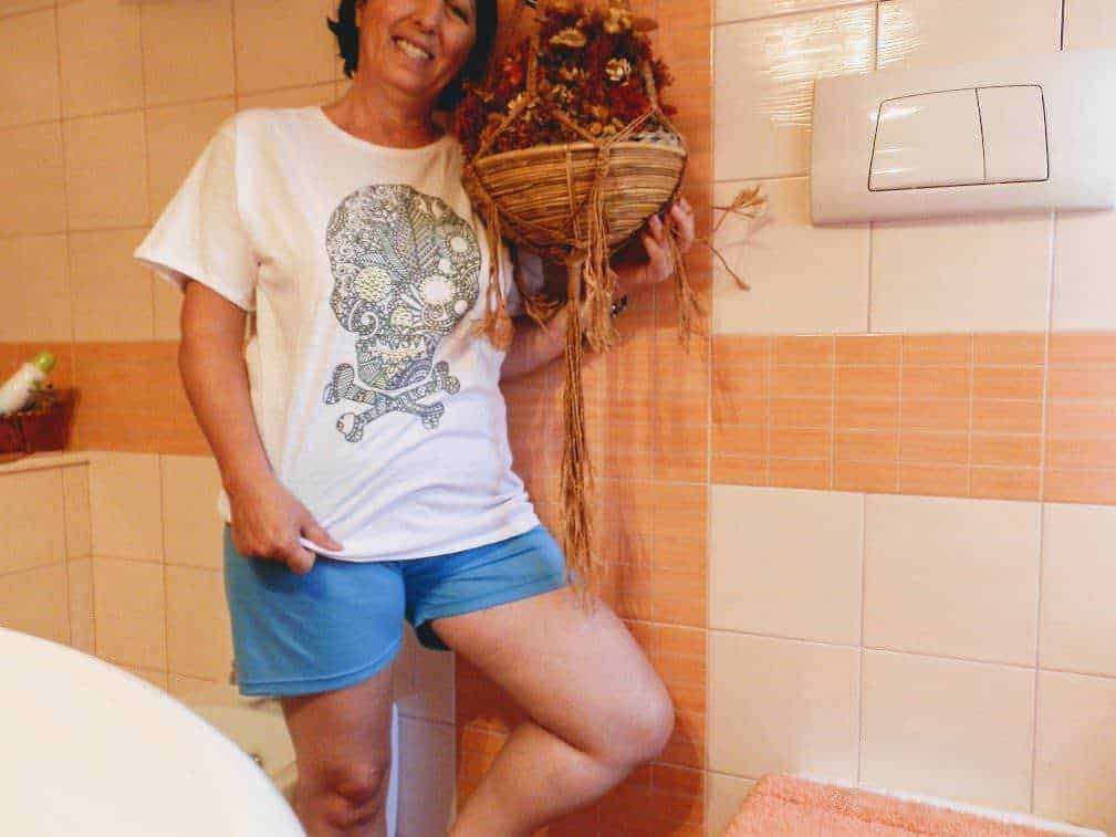 Femminile con maglietta con stampa skull e sedia a rotelle con decorazioni natalizie, in ambiente bagno con pareti in piastrelle arancioni e oggetti decorativi, in un contesto di vita quotidiana.