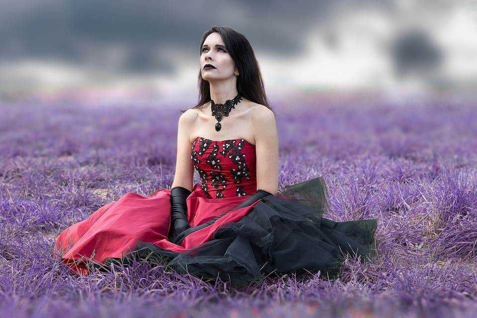 Elegant woman in gothic style dress sitting in purple lavender field, dark makeup, black lace necklace, and long black gloves, evocative of romantic and mysterious aesthetics.