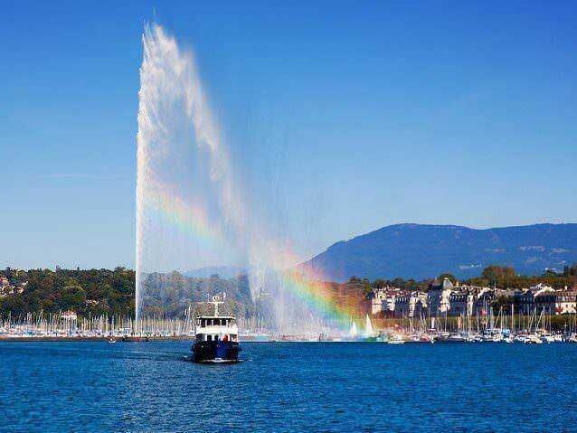 Jet d'eau di Ginevra con arcobaleno sul lago, panorama della città e delle montagne sullo sfondo, attrazione turistica iconica in Svizzera.