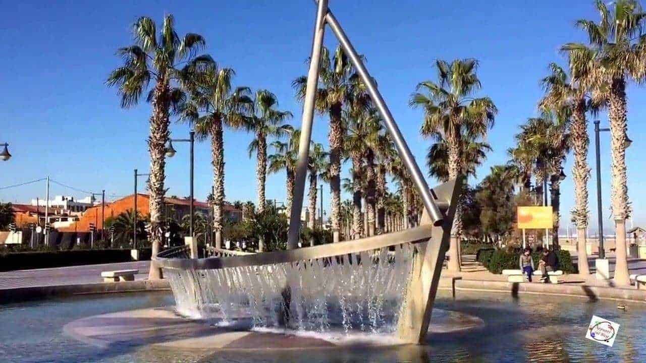 Fioriera moderna in metallo con fontana di acqua al centro, palme e area pedonale in背景 in una città costiera soleggiata, perfetta per rilassarsi e passeggiare.