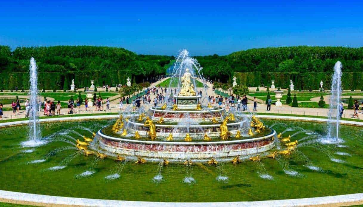 Fontana decorativa con sculture dorate nel parco di un palazzo storico, con molti visitatori e un ampio giardino ben curato sotto un cielo blu brillante.