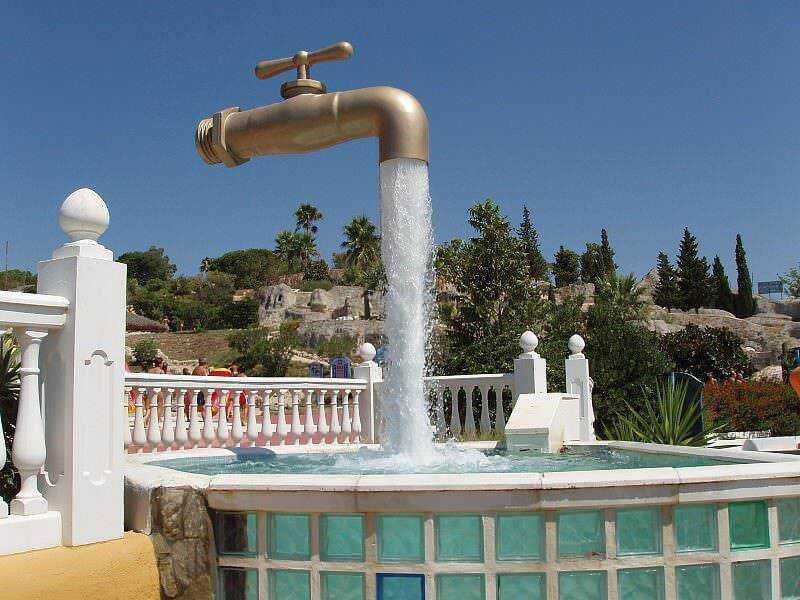 Tubo di sciacquone che funge da fontana decorativa in un giardino con piscine, ornamenti bianchi e paesaggio verde, atmosfera estiva soleggiata.
