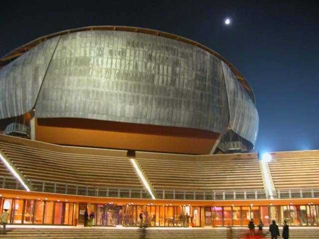 Un'immagine del Forum di Assago di sera, con luci decorative e pubblico all'ingresso, sotto un cielo notturno con luna, rappresenta un moderno centro congressi e concerti in Lombardia.