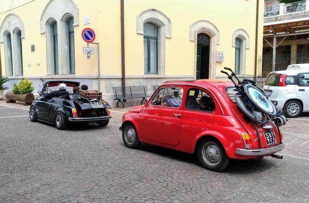 Vintage Fiat 500 parked on street, carrying bicycles on the back, in front of classic Italian building, showcasing retro automotive style and urban ambiance.