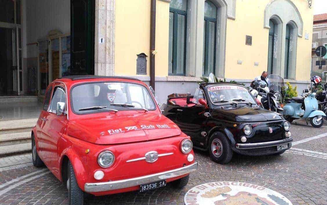 Fiat 500 vintage rossa parcheggiata in strada tra scooter e motociclette, ambientazione urbana storica