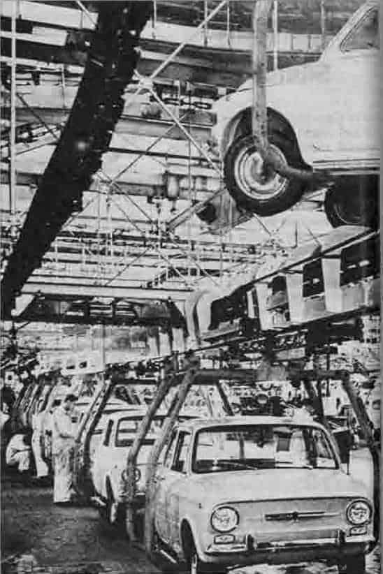 Officina auto con linee di produzione e veicoli in fase di assemblaggio, rappresentando l'industria automobilistica in un contesto storico industriale.