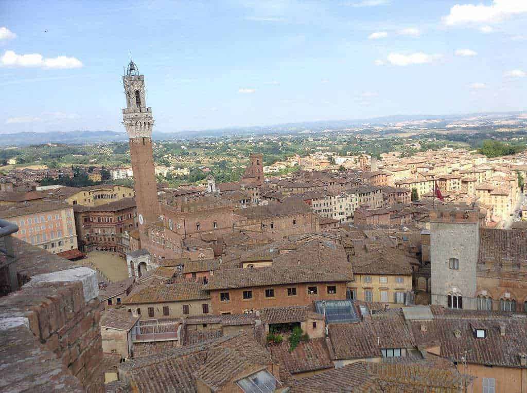 Torri e tetti di una città medievale in Toscana, vista panoramica su Pisa con Torre Pendente, cielo blu e paesaggio rurale. Immagine suggestiva di un centro storico italiano ricco di storia.