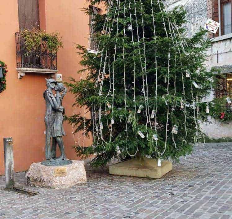Albero di Natale decorato con luci e biglietti in una piazza di paese, accanto a statua di due persone abbracciate in un contesto rurale, con case in pietra e pavimentazione in cobblestone, atmosfera natalizia e tradizionale.