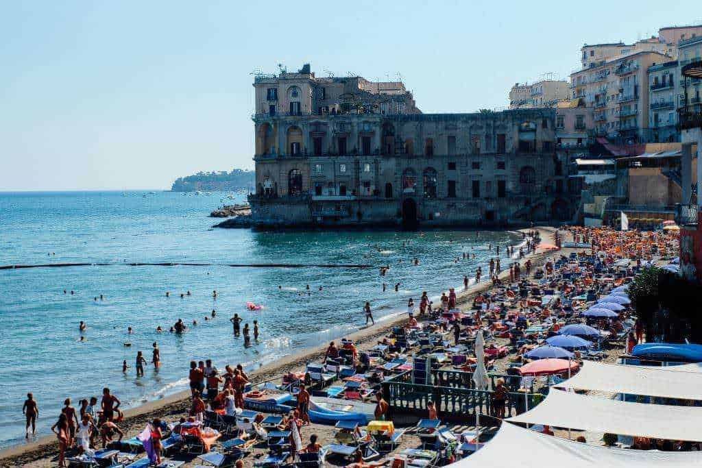 Spiaggia affollata con ombrelloni, persone che nuotano e un edificio storico sul mare a Napoli, Italia, durante una calda giornata estiva, perfetta per vacanze al mare e relax.